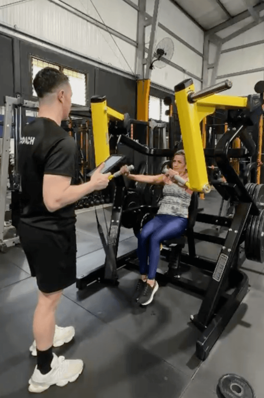 A personal trainer guides a client using a chest press machine in a gym setting.