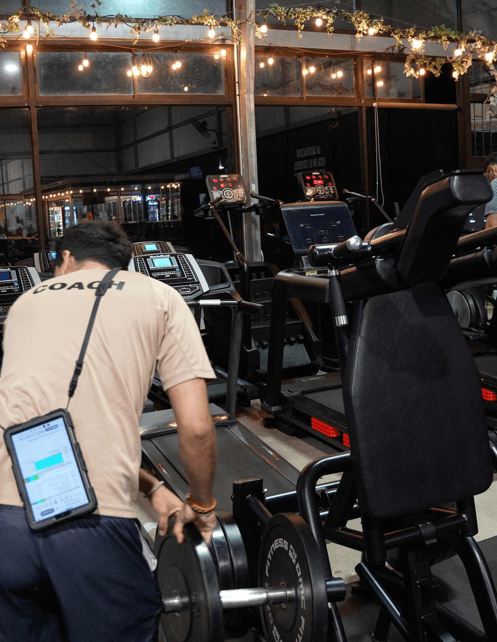 Gym interior with a coach adjusting weights, treadmills in the background, and decorative lights above.