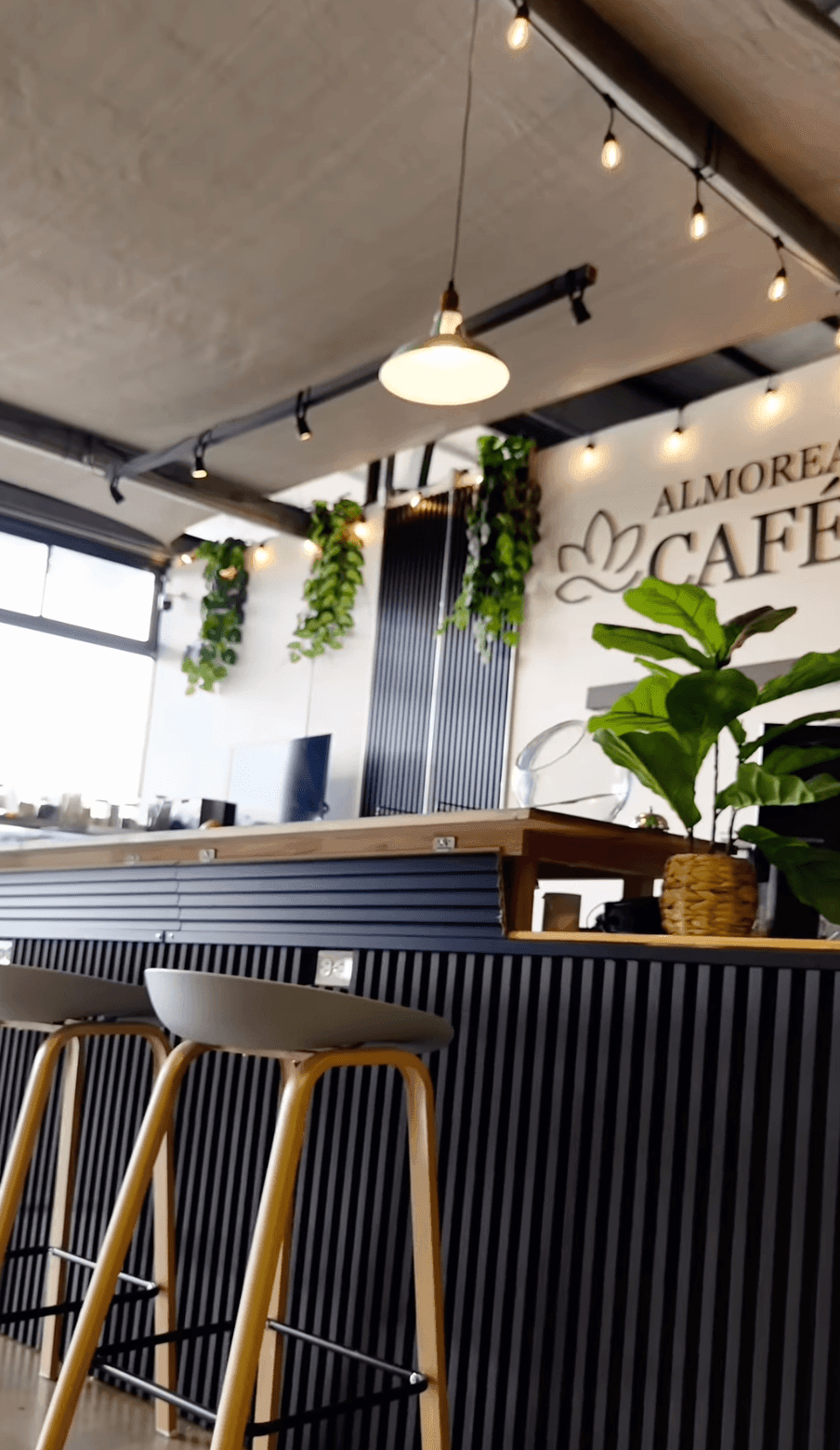 Modern cafe interior with plants, hanging lights, and wooden bar stools at a counter.